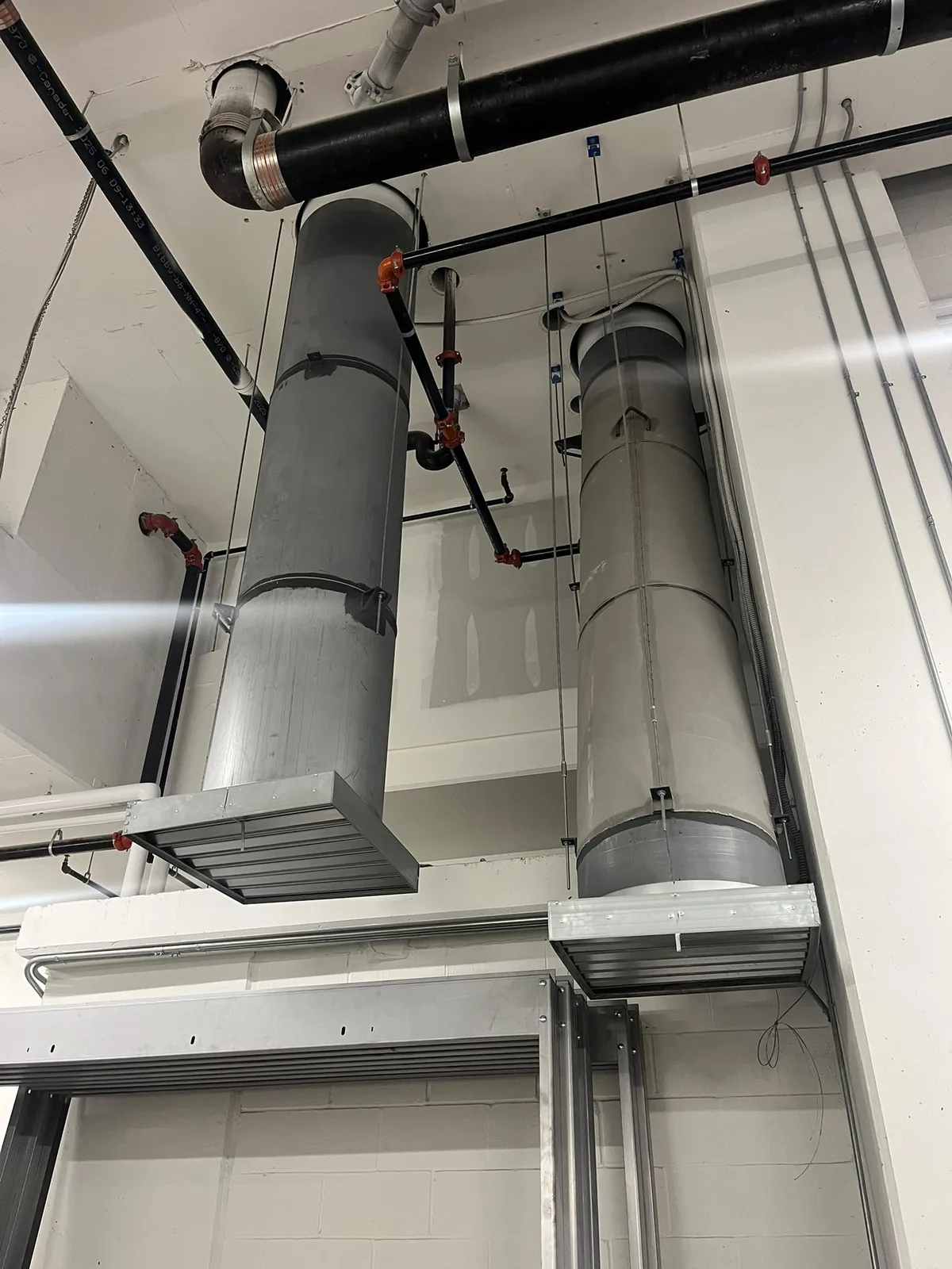 Twin grey cylindrical duct risers ending in fire dampers against a block wall with black piping