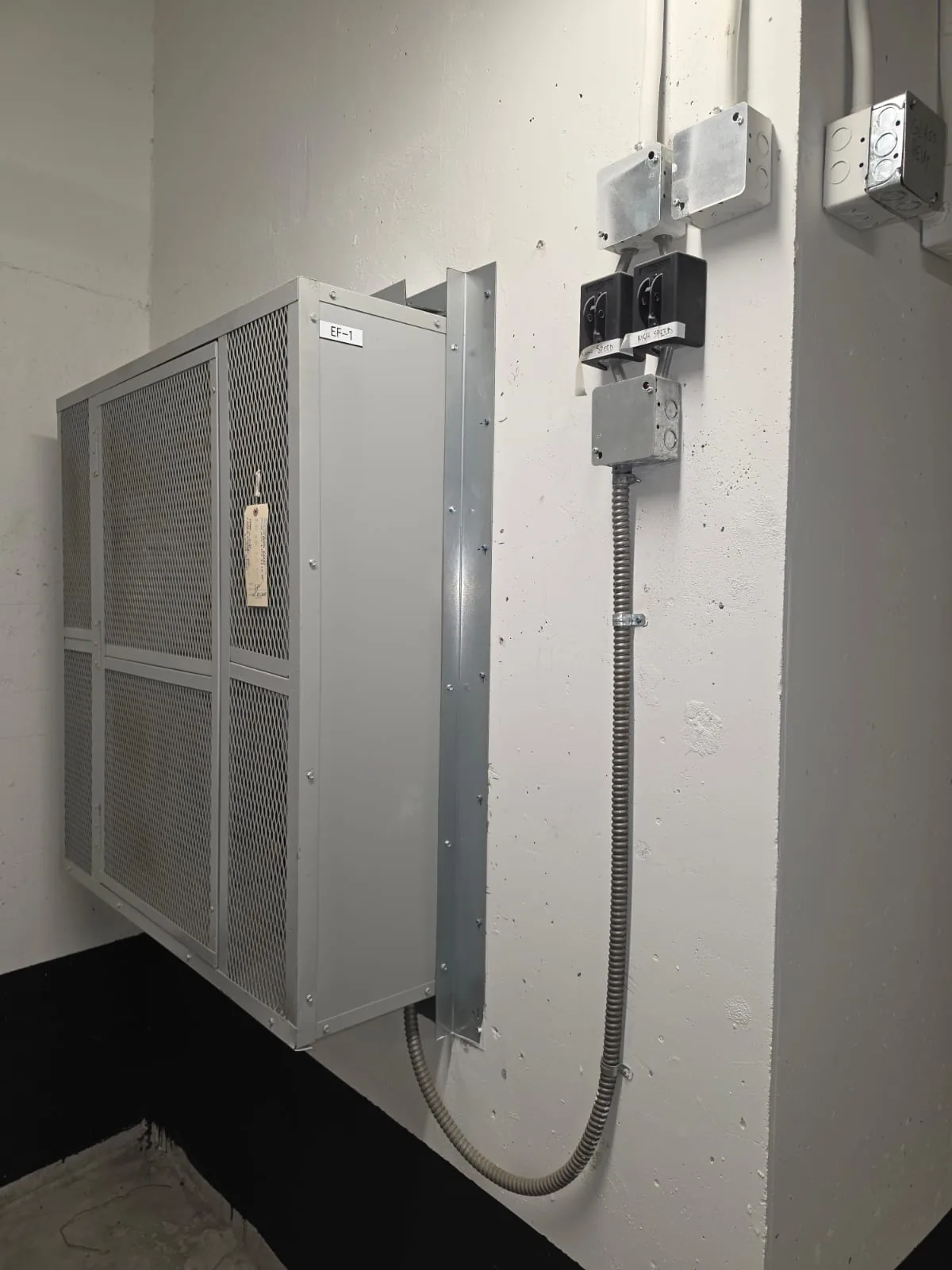 Wall-mounted EF-1 exhaust fan cabinet with mesh grille and flexible conduit on a block wall