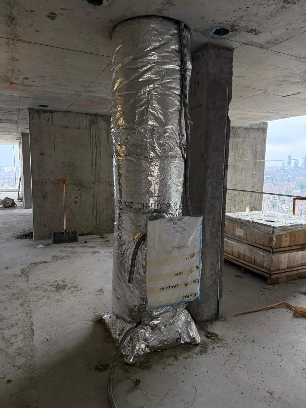 Foil-wrapped vertical duct riser beside a concrete column on an unfinished high-rise floor