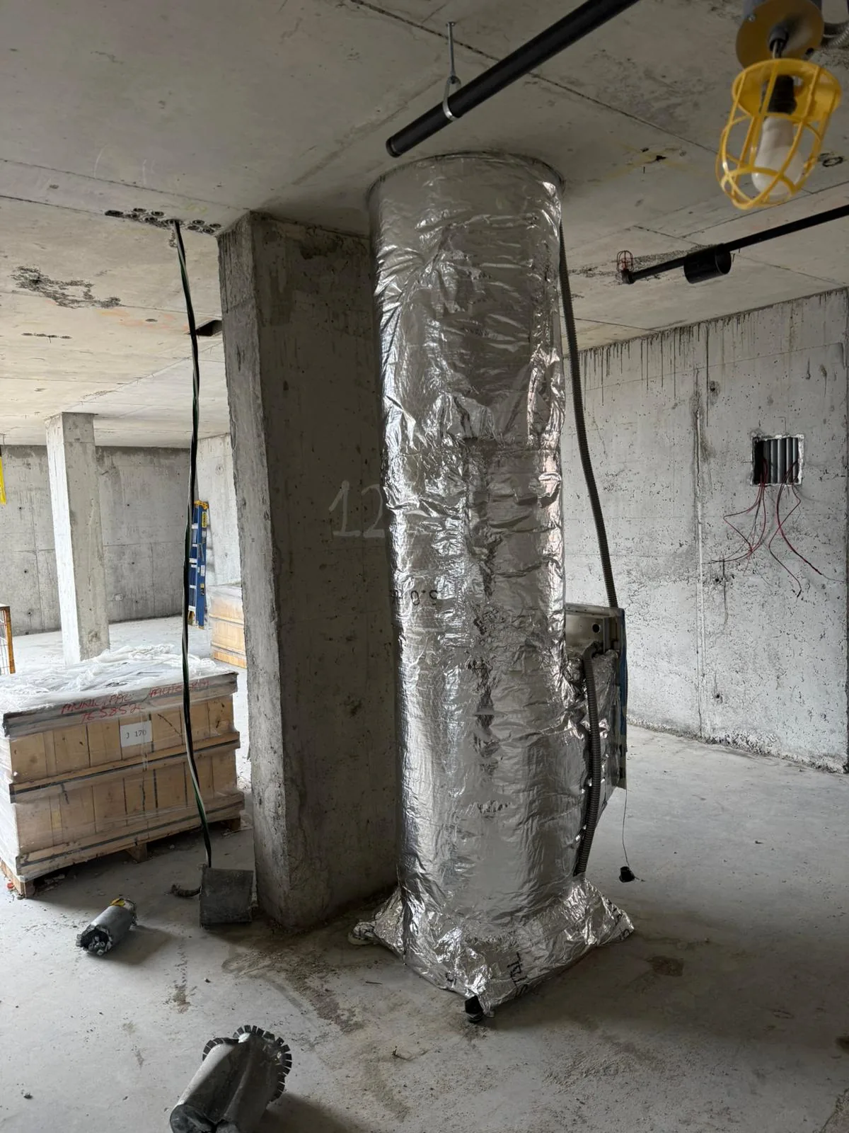 Foil-wrapped vertical duct riser against a concrete column under a caged construction light