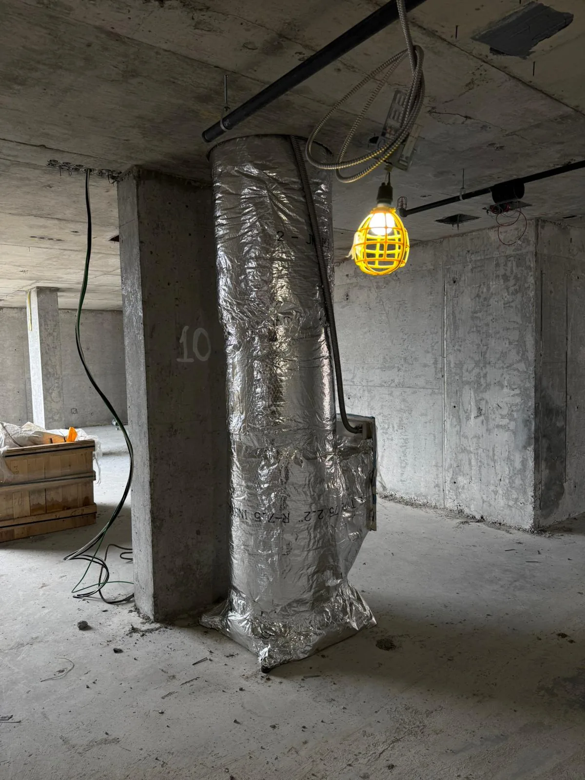 Foil-wrapped duct riser beside a numbered concrete column with a caged work light overhead
