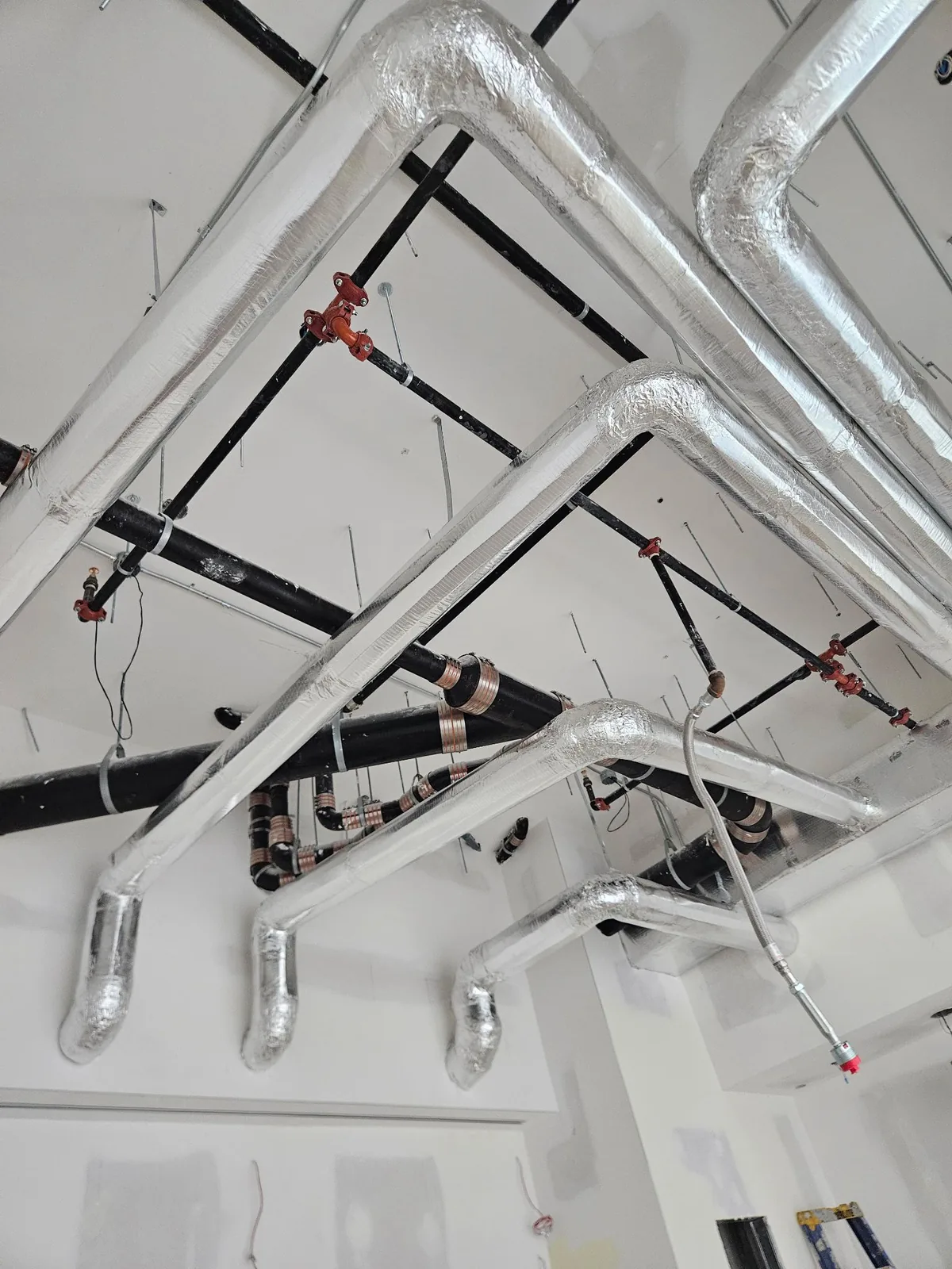 Foil-wrapped insulated pipe runs with swept elbows across a white mechanical room ceiling