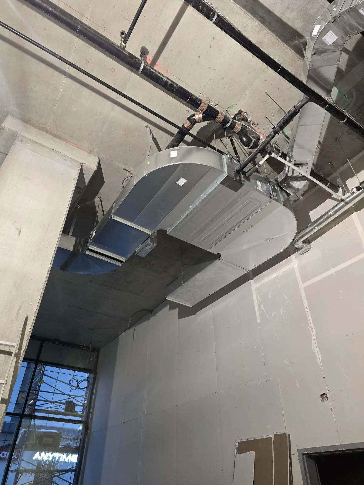 Oval duct-mounted air handling unit suspended beneath black piping above drywall walls