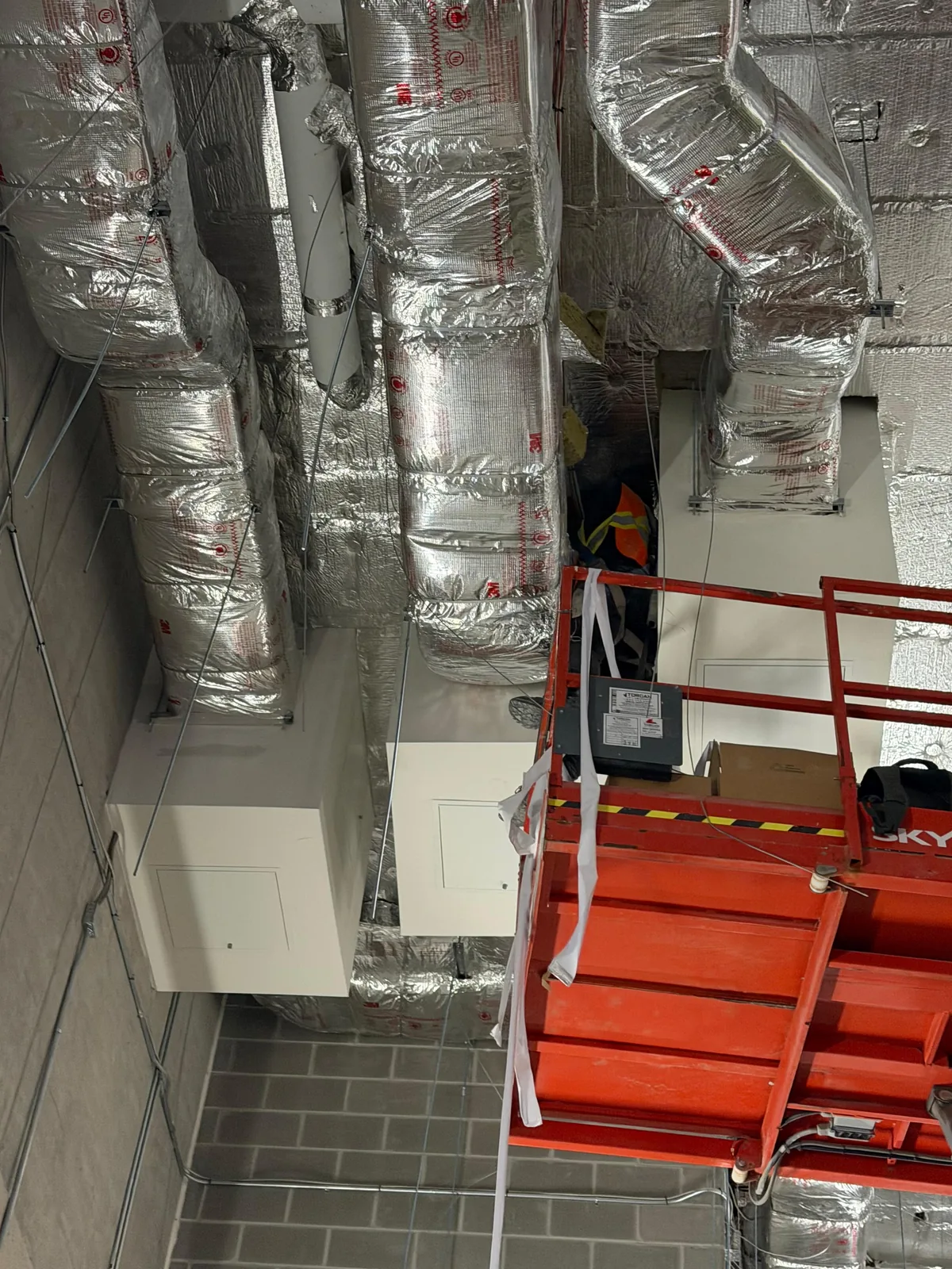 Foil-wrapped flexible ducts dropping to grey equipment cabinets above an orange scissor lift